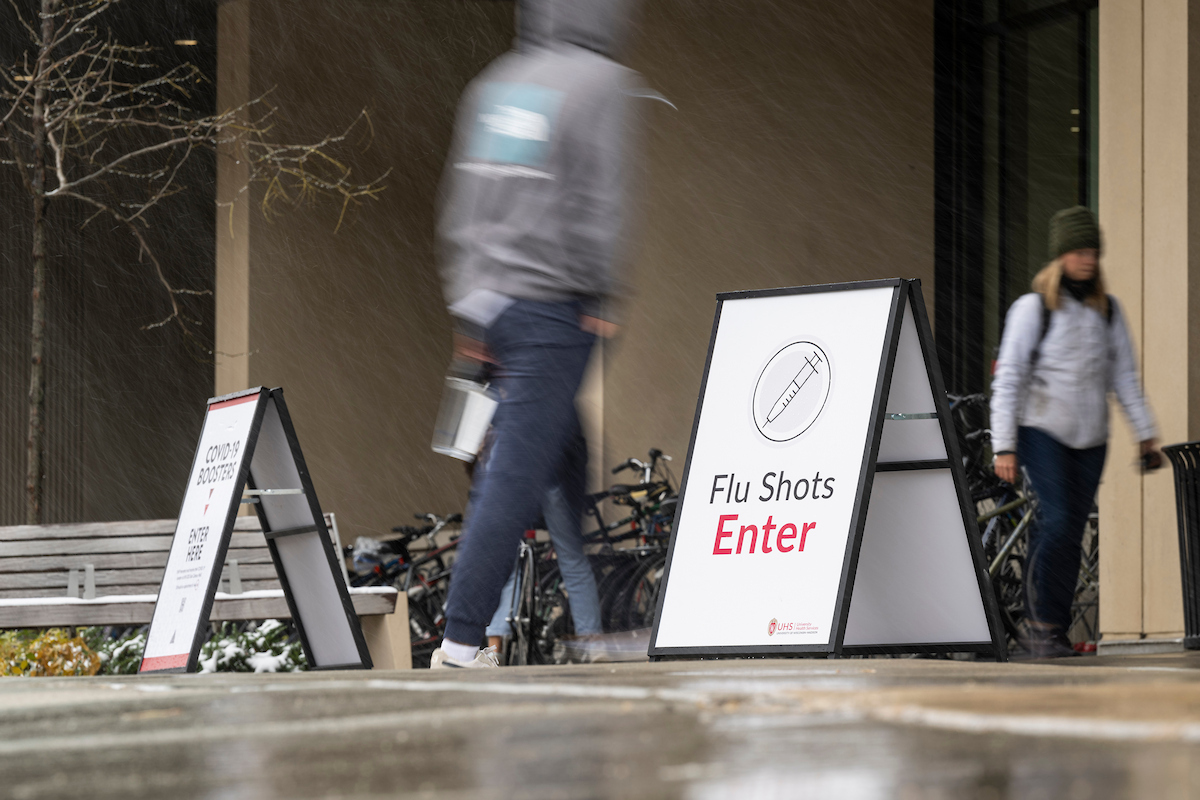 Pedestrians walk outdoors past signage advertising flu shots as part a COVID-19 booster and influenza vaccination clinic held at the Nicholas Recreation Center (the Nick) at the University of Wisconsin–Madison during a wet and snowy day on Nov. 15, 2022. (Photo by Jeff Miller / UW-Madison)